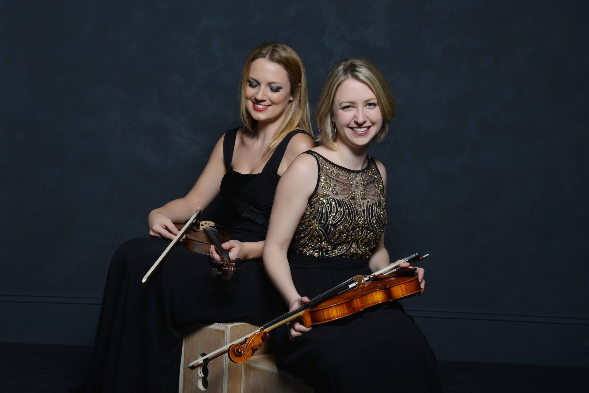 Lyrae duo Lianna Jeffery & Jennifer Maslin sat smiling on a box each holding a violin